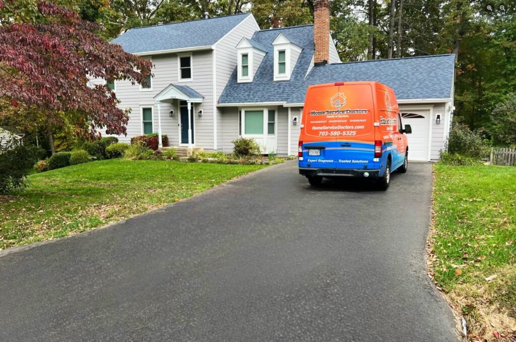 Service van parked at top of a home's driveway, in front of the garage. Home is large white colonial style with trees in background.